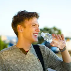 Un jeune homme souriant boit de l'eau d'une bouteille en plastique par une journée ensoleillée.