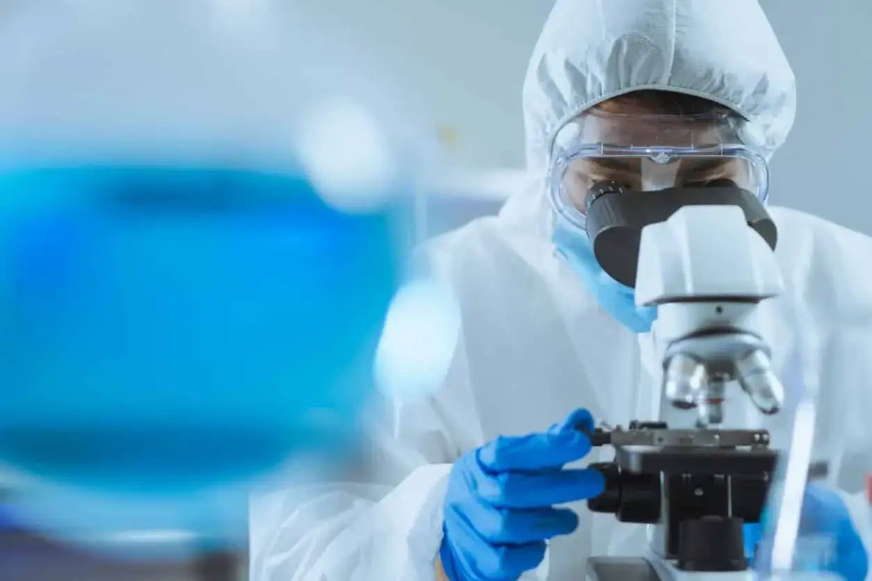 Chercheuse en blouse de protection examinant une goutte d'eau du robinet au microscope, avec un liquide bleu en arrière-plan.
