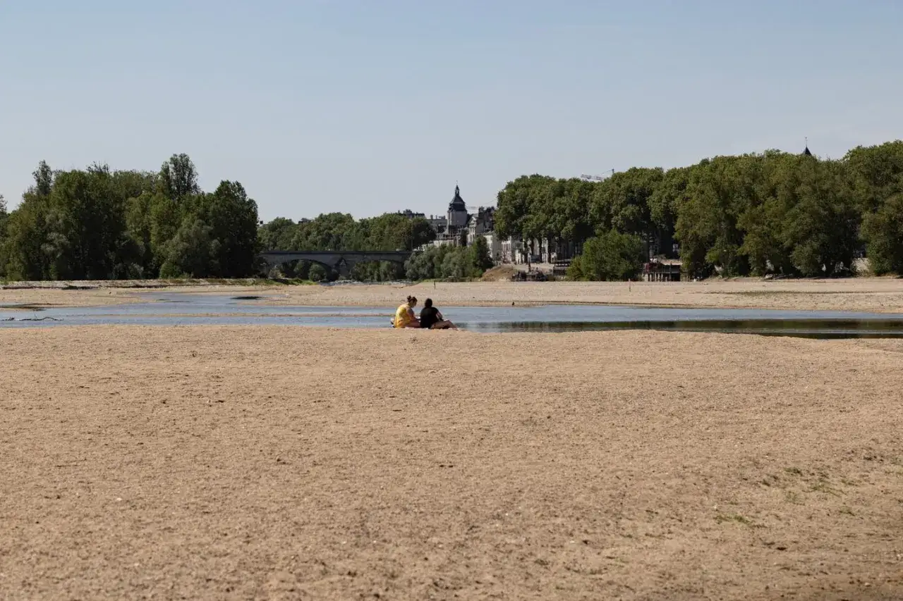 Deux personnes assises sur une plage de sable sec, témoignant du manque d'eau. Au loin, un pont et des arbres bordent un mince filet d'eau.