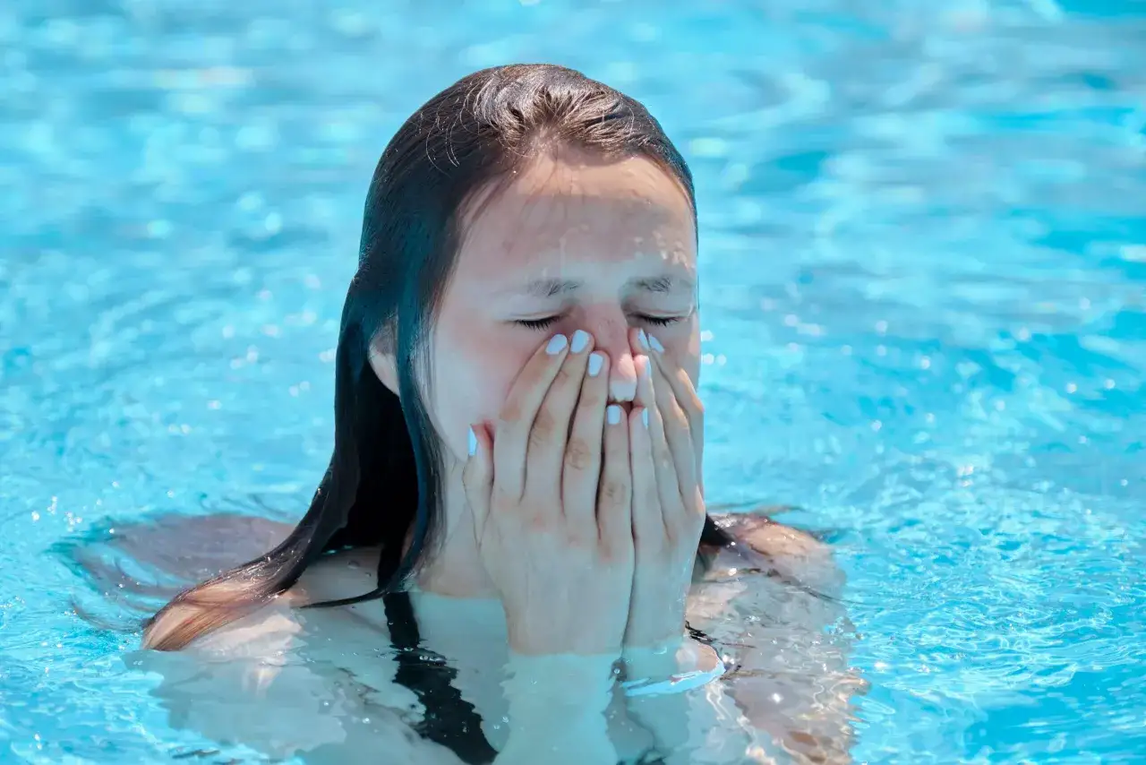 Jeune fille dans une piscine, le visage couvert par ses mains, possiblement à cause d'une allergie au chlore.